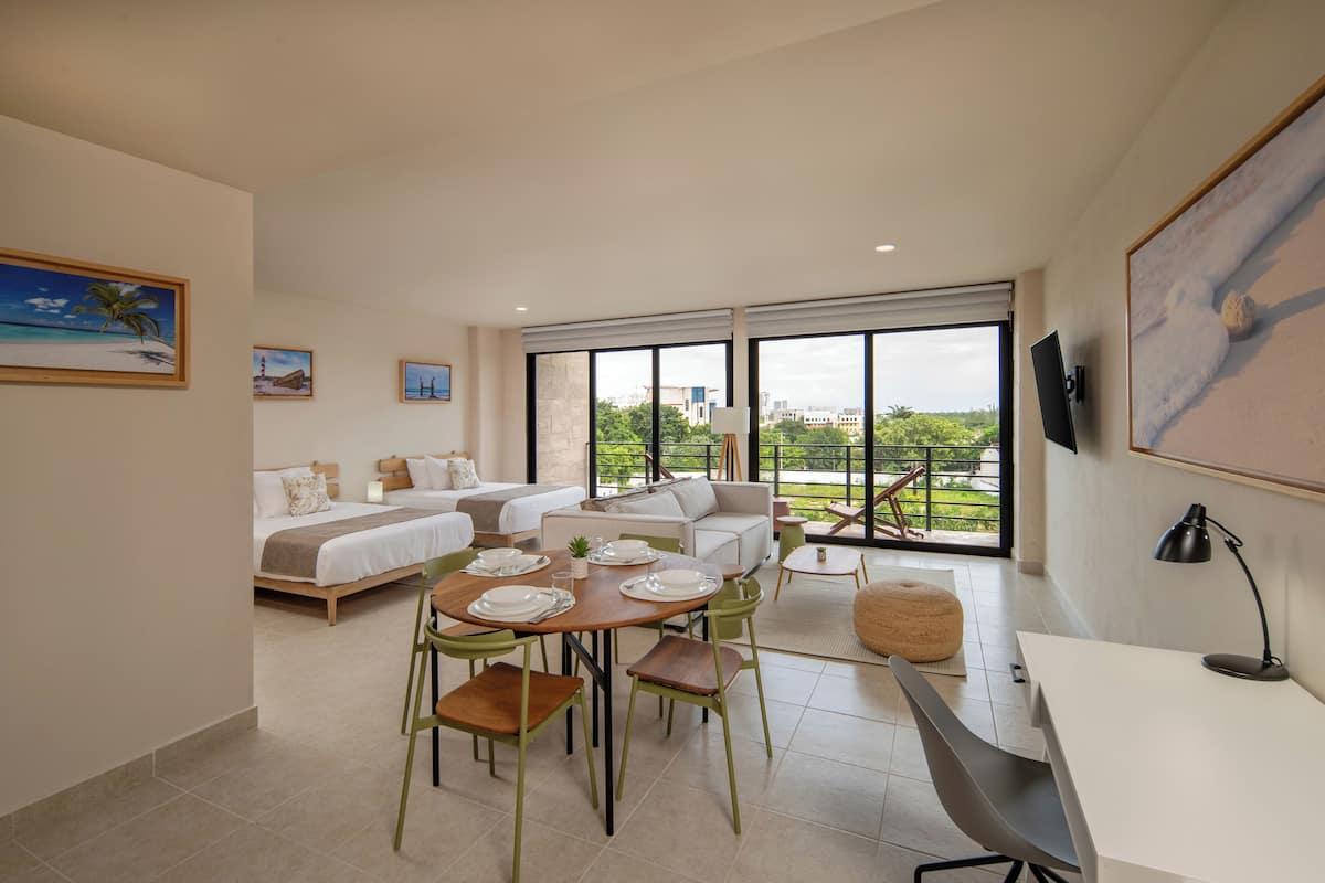 Habitación de hotel moderna con cama, sofá, mesa de comedor y sillas. Hay un televisor montado en la pared y una ventana con vista a zonas verdes.