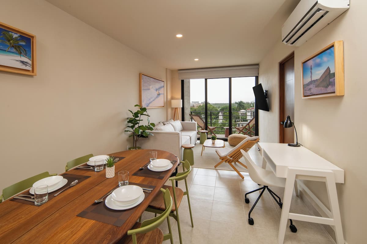 Un comedor con una mesa de madera dispuesta para cuatro personas, un sofá, un escritorio y un televisor montado en la pared.