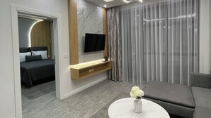 A modern living room with a flat-screen TV, a round coffee table with a vase, a grey sofa, and a bed in the adjacent room.