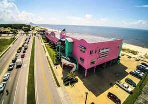 Un bâtiment rose avec une enseigne indiquant « Sharknado's », situé sur une route côtière bordée de voitures garées, avec vue sur l'océan.