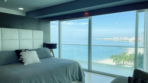A modern bedroom with a large bed, a nightstand, and a view of the ocean. - Lujoso Peninsula T3 Frente al Mar (Puerto Vallarta)