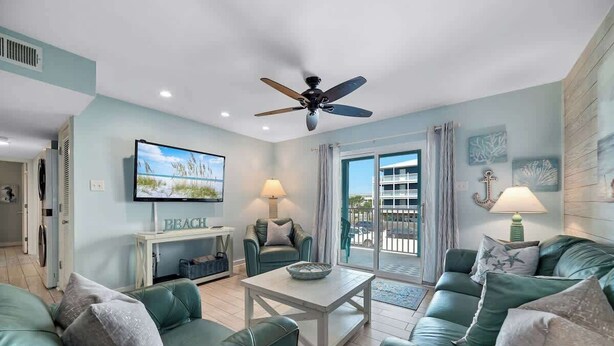 A living room with teal sofas, a ceiling fan, a TV, and a balcony with a view. - Affordable updated 2BR family retreat with pool (Gulf Shores)