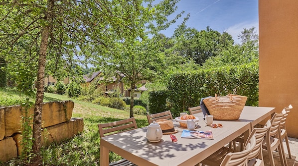 An outdoor dining area with a table, chairs, a basket, a hat, a teapot, and a magazine.