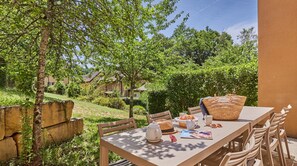 Un espace de restauration en plein air avec une table, des chaises, un panier, un chapeau, une théière et un magazine.