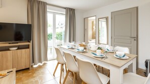 A modern dining area with a long table set for four, a television on a wooden stand, and a sliding glass door leading to an outdoor area.