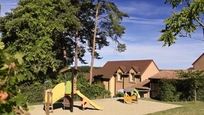 A playground with a slide and climbing structures, surrounded by trees and a building with a tiled roof.
