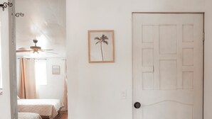 Une pièce avec un plancher en bois, une porte blanche dotée d’un trou de serrure, un tableau encadré représentant un palmier et un ventilateur de plafond.