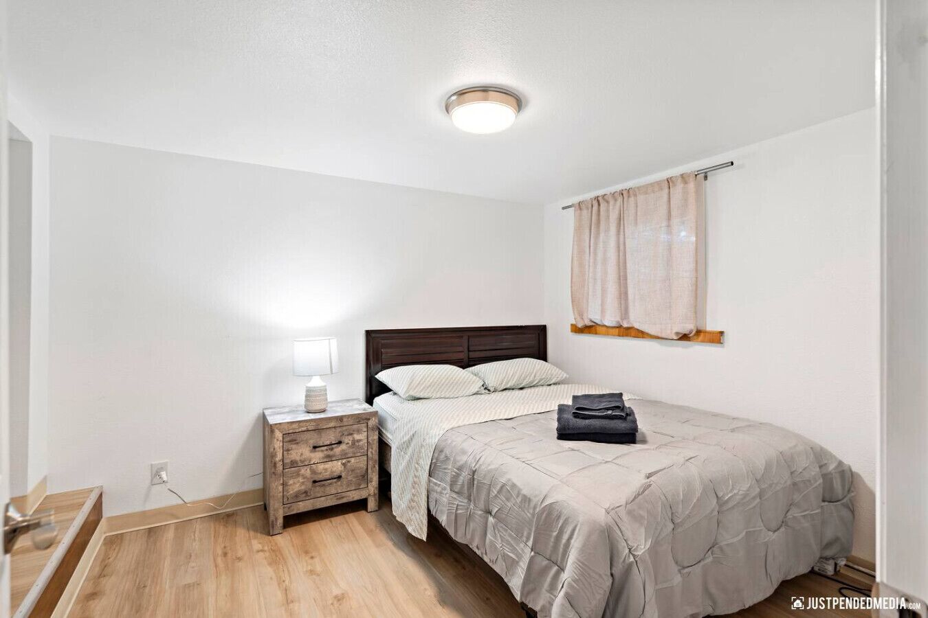 A bedroom with a wooden nightstand, a bed with a beige quilt, a window with beige curtains, and a ceiling light.