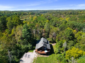 An aerial view of a cabin surrounded by dense forest, with a deck and outdoor seating area.