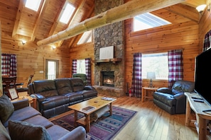 A cozy cabin interior with a stone fireplace, leather sofas, and a wooden ceiling.