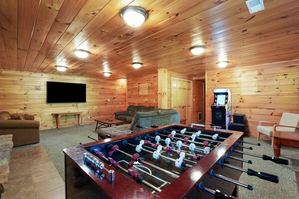 A cozy room with wooden walls, a foosball table, a sofa, a television, and a small gaming machine.