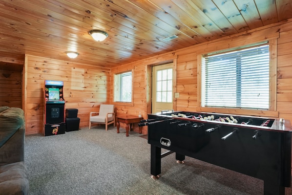 A cozy room with wooden walls and ceiling, a foosball table, a small table with a chair, and a foosball machine.