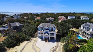 A large, multi-story house with a prominent balcony and a swimming pool, surrounded by dense trees and a view of the ocean. - Oceanside, Private Pool, Hot Tub, WC960, Blue Heron (Corolla)