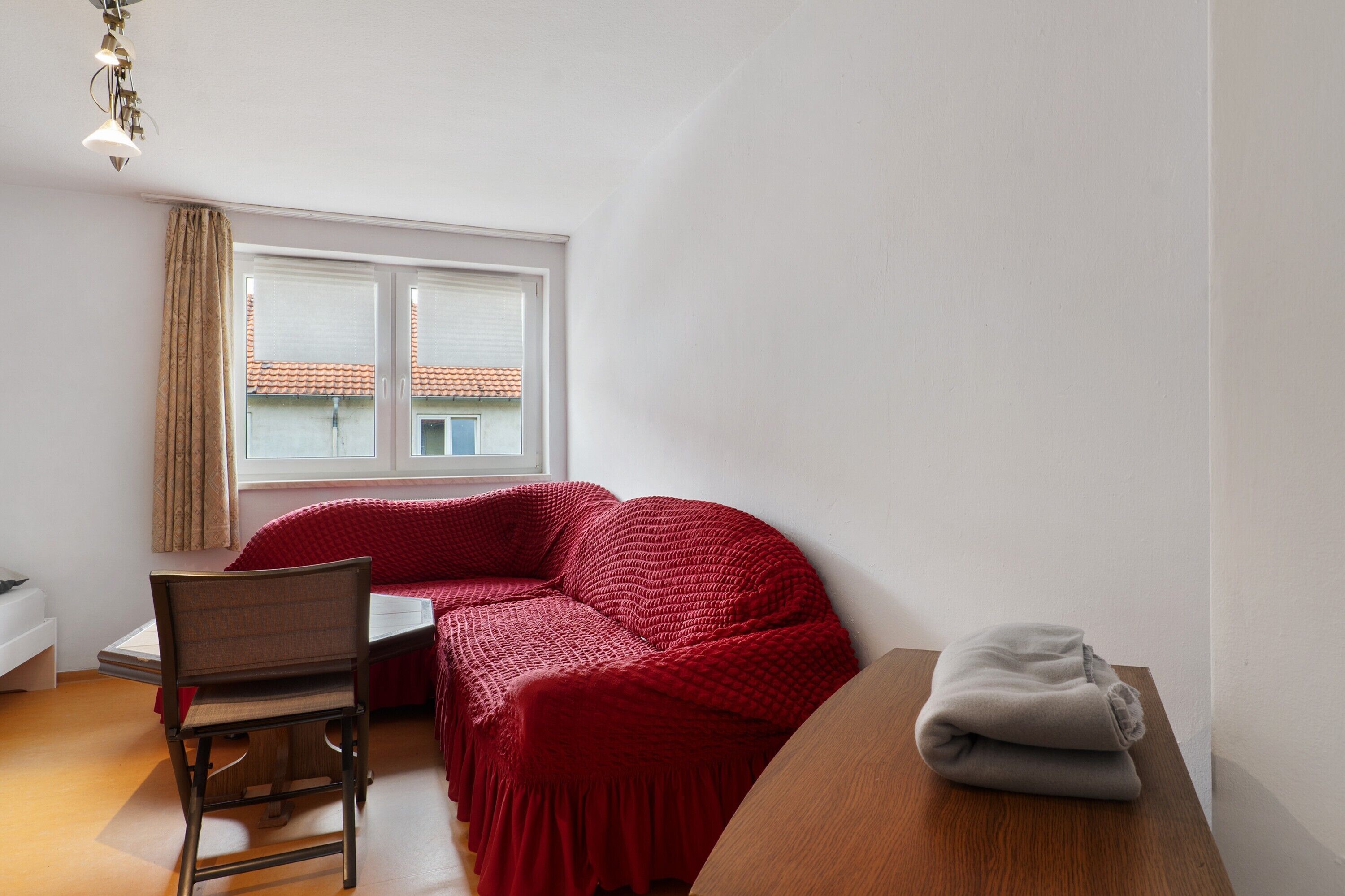 Une pièce avec un canapé rouge, une table en bois, une chaise et une fenêtre avec des stores.