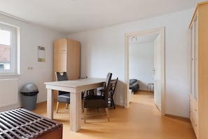 Una habitación con una mesa de madera, dos sillas, un cubo de basura, un radiador y una puerta que da a otra estancia.