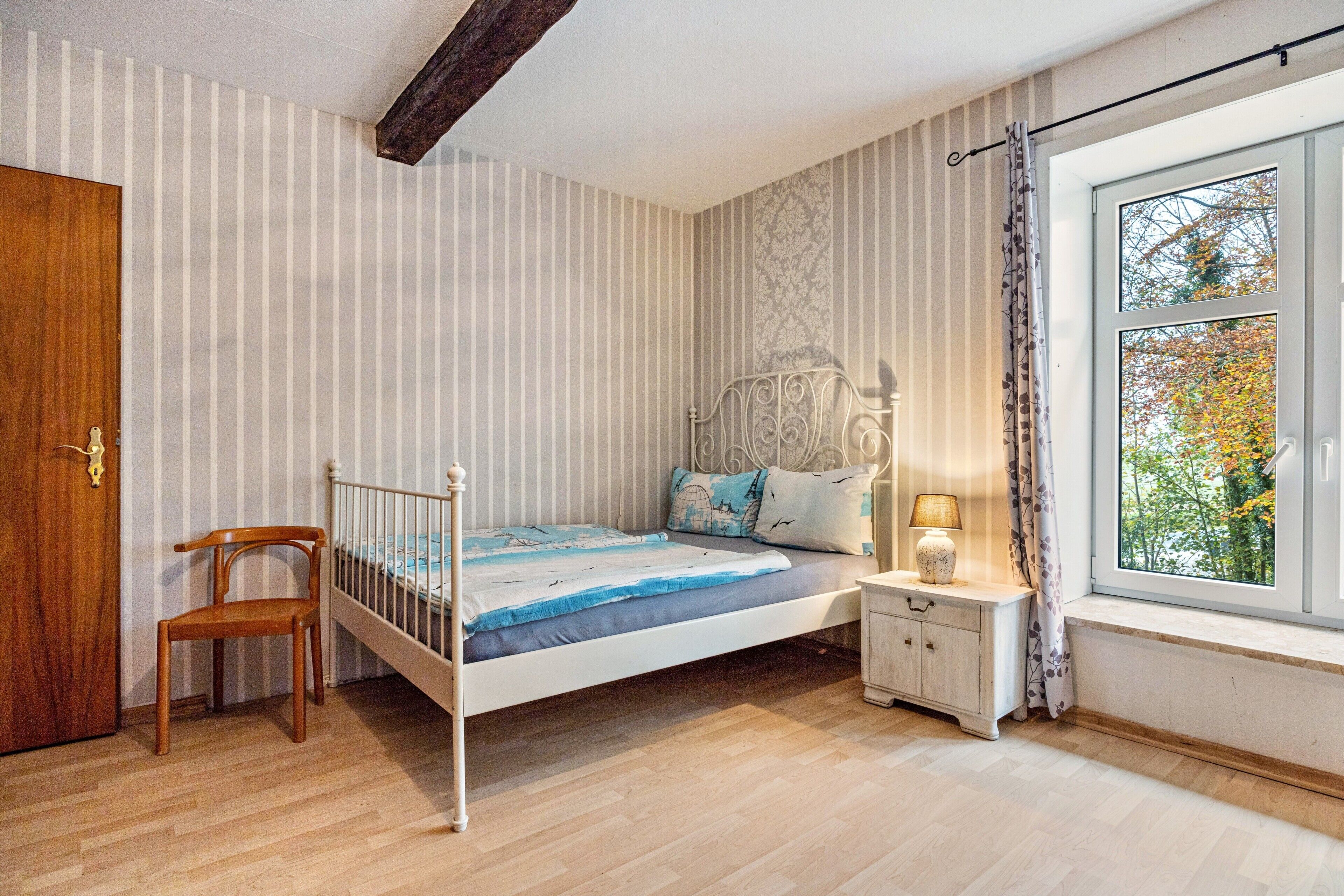 A bedroom with a bed, a wooden chair, a nightstand, and a window with a view of trees.