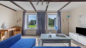 Una sala de estar con un sofá, una mesa, un televisor y ventanas grandes con cortinas.