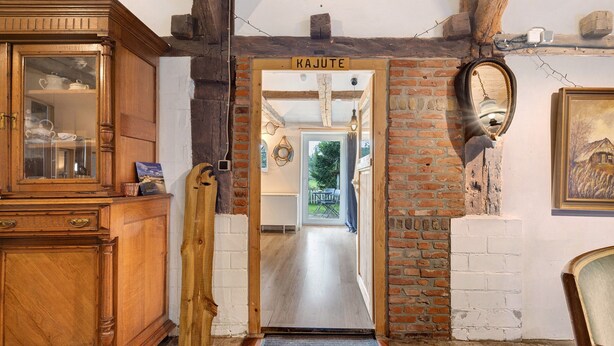 Un armario de madera con puertas de cristal, una puerta de madera con la etiqueta 'KAJUTE', y una pared de ladrillo con un espejo colgado.