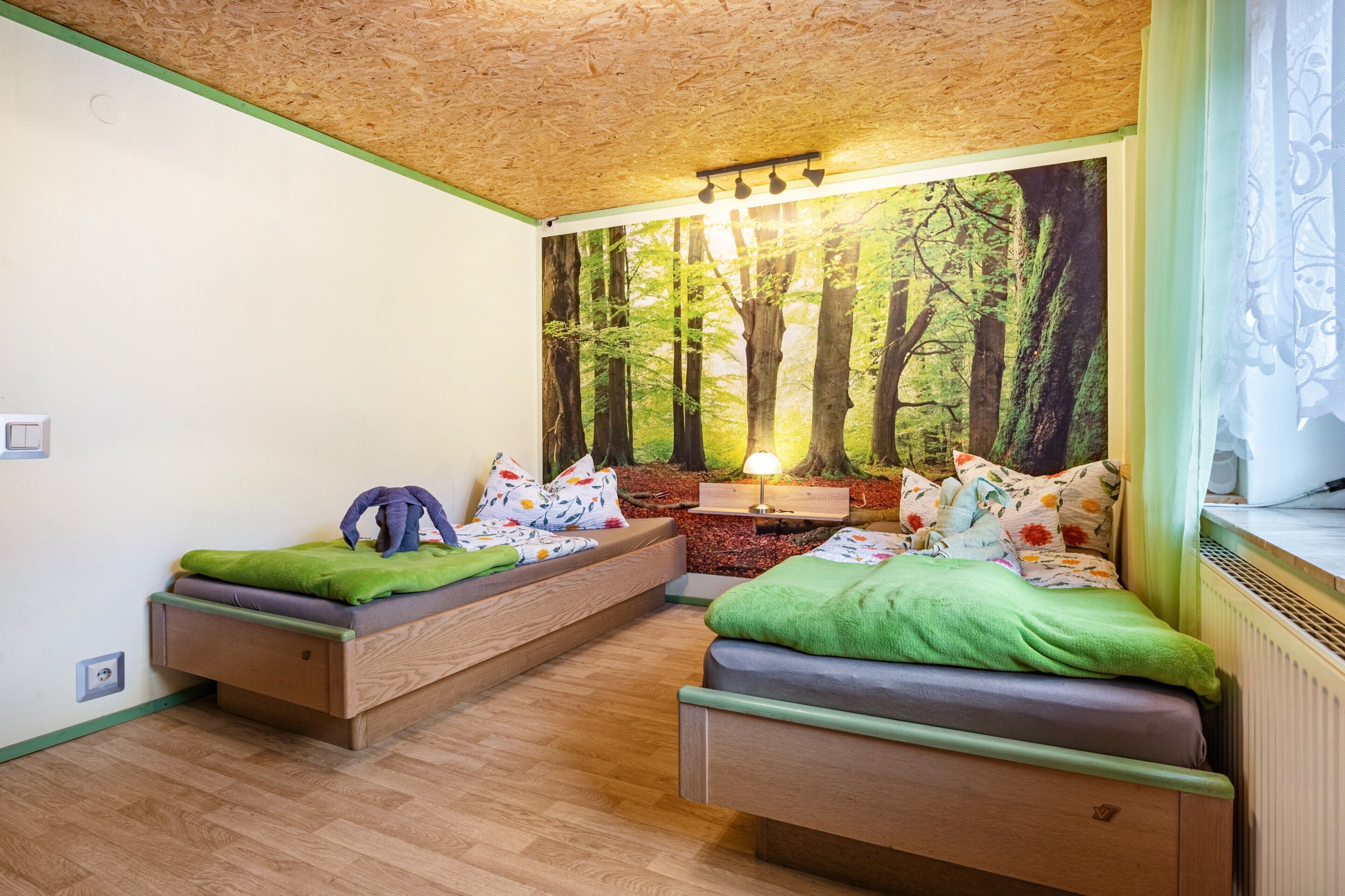 Quarto com duas camas, um mural de uma floresta, uma luminária e uma janela com cortinas.
