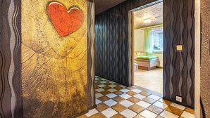 A hallway with a checkered floor, a wooden wall with a heart design, and a room with a bed and curtains in the background.