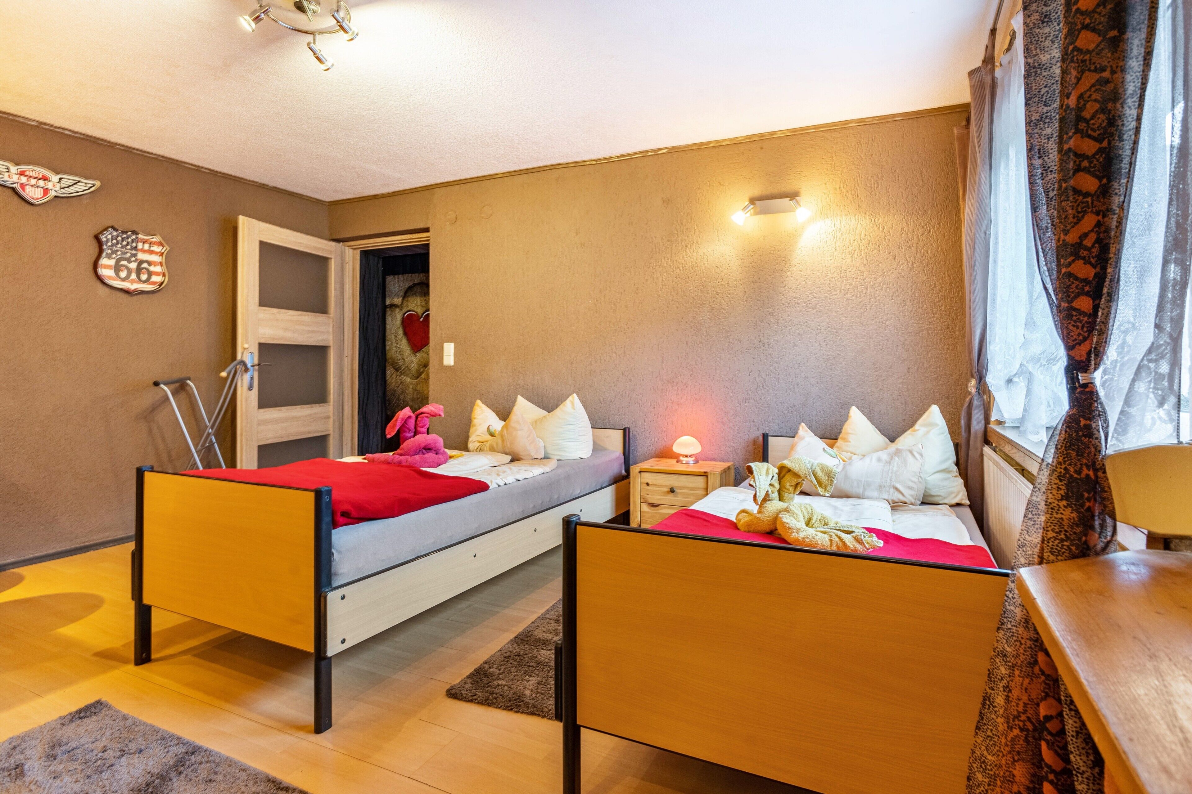 A bedroom with two beds, a nightstand, a lamp, a window with curtains, and a door leading to another room.