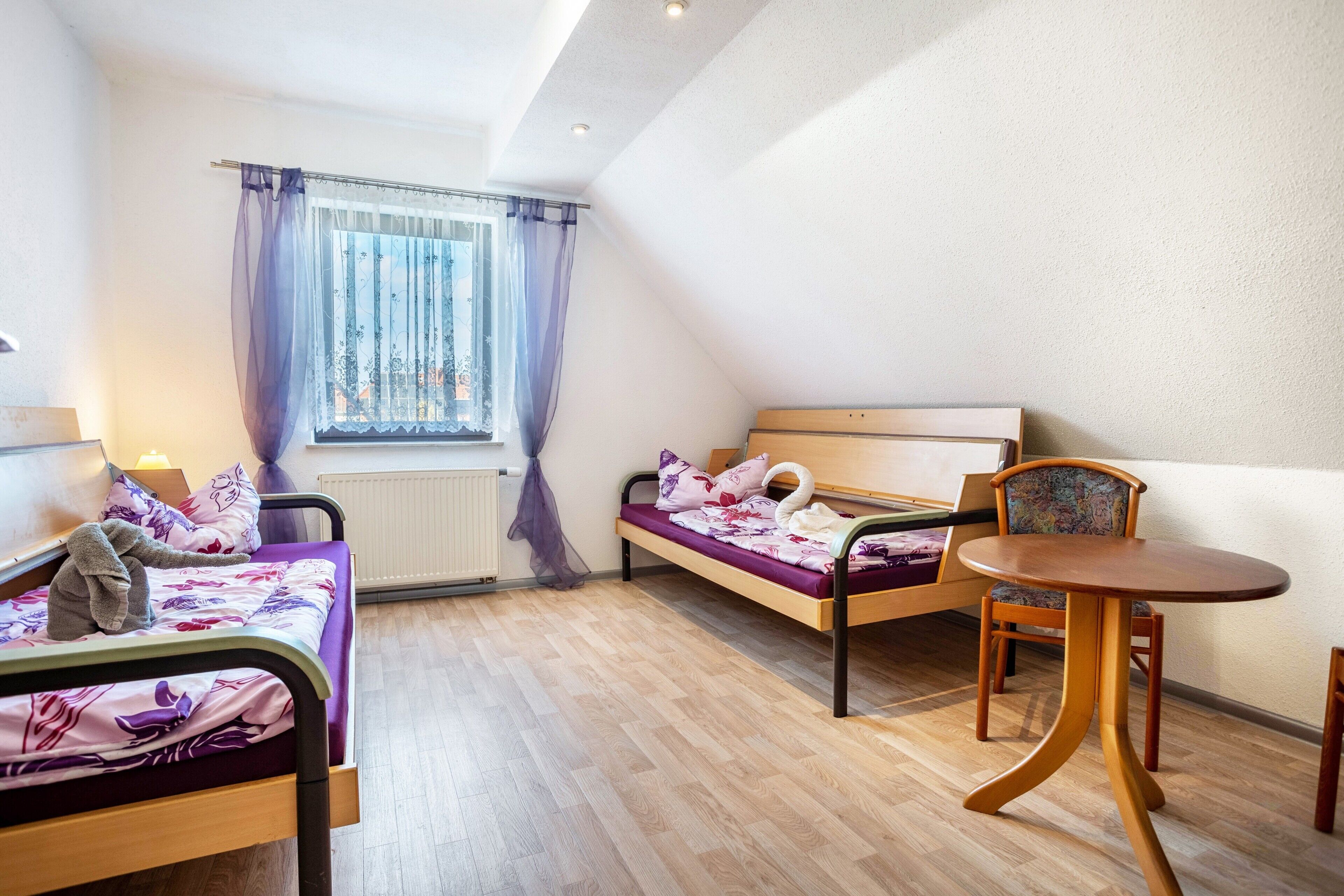 A small, well-lit room with two beds, a round wooden table, and a window with curtains.