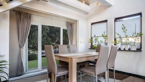 Un espace repas comprenant une table et des chaises, de grandes fenêtres avec des rideaux et des plantes en pot.