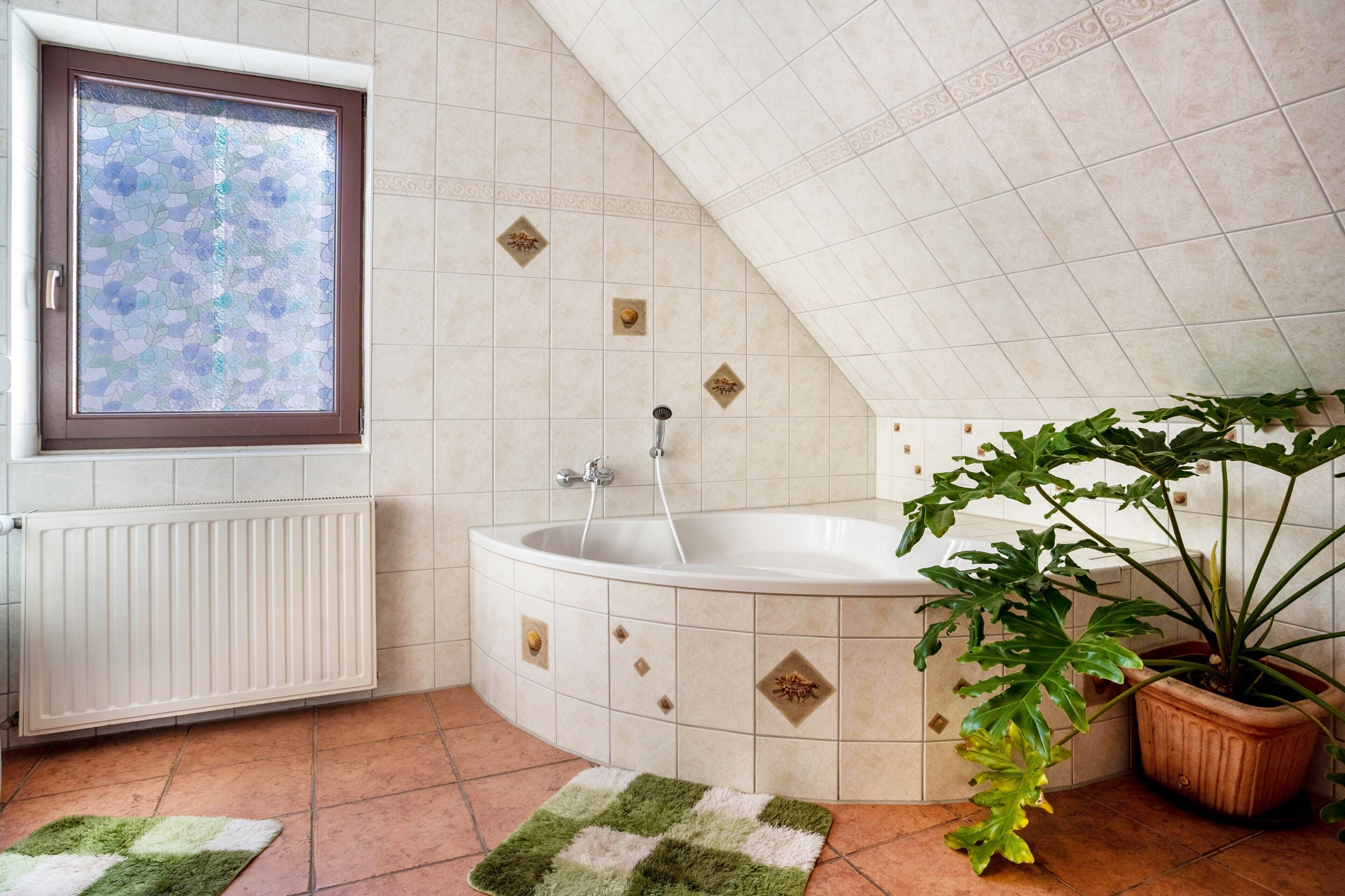 Un bagno con una vasca freestanding, una finestra con un pannello decorativo in vetro, un termosifone, una pianta in vaso e un tappeto verde e bianco.