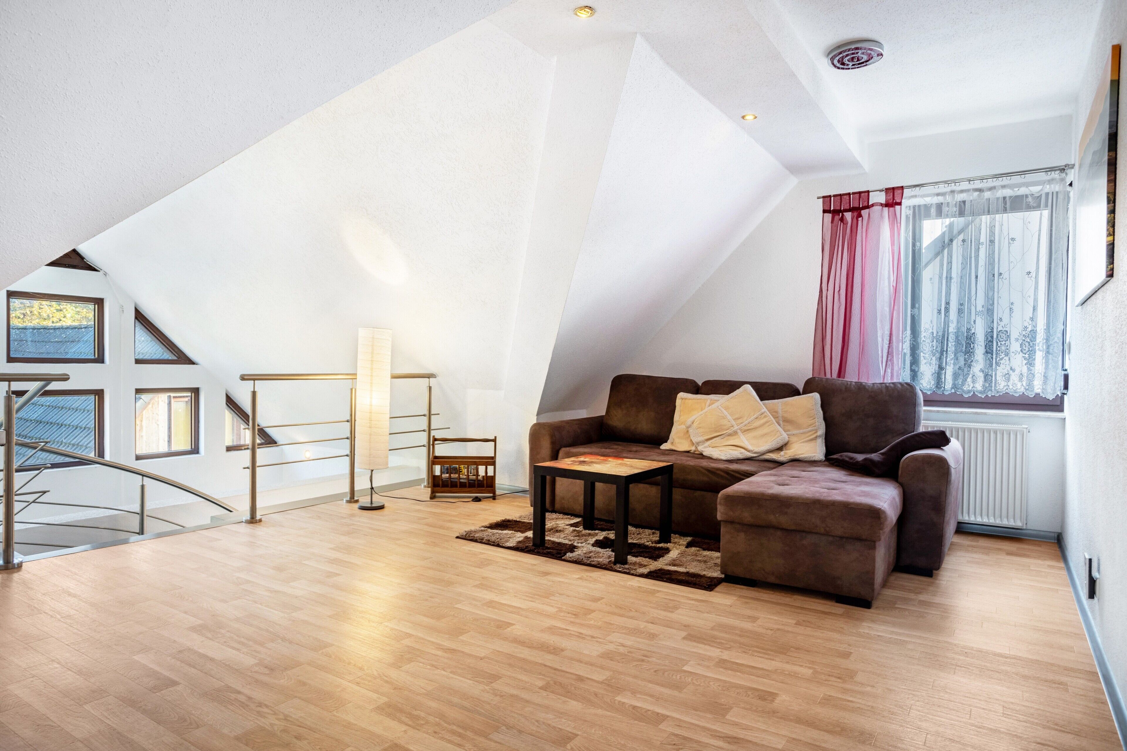 Une pièce de style loft avec un plafond en pente, un canapé marron, une petite table et une fenêtre ornée de rideaux légers.
