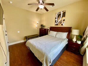 A bedroom with a bed, wooden floor, ceiling fan, bedside table with lamp, and artwork on the wall.