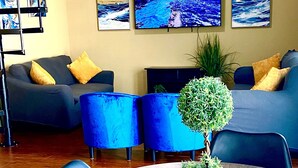 A living room with a round wooden table, blue chairs, a grey sofa, and a ceiling fan. There are three framed pictures on the wall.