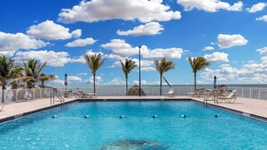 A rectangular swimming pool with clear blue water, surrounded by palm trees and lounge chairs, with a view of the ocean and a clear sky. - Ocean Isles Seaside Oasis (Marathon)