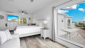 A bedroom with a bed, a nightstand, a TV, a ceiling fan, and a large window with a view of a white building and palm trees. - Ocean Isles Seaside Oasis (Marathon)