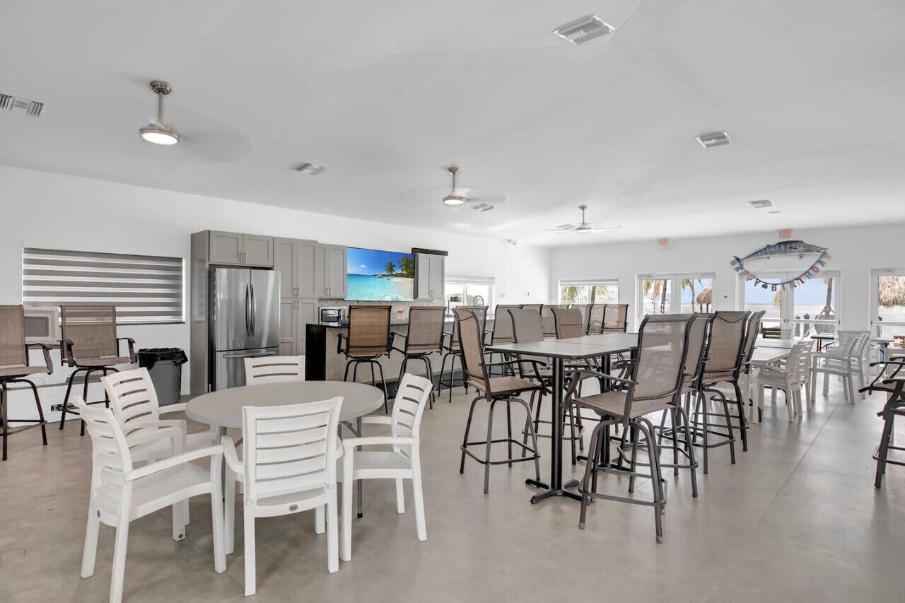 Une cuisine spacieuse équipée d’appareils modernes, un coin repas avec plusieurs tables et chaises, et de grandes fenêtres offrant une vue sur l’extérieur.