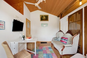 Ein gemütliches Zimmer mit Holzbalkendecke, einem an der Wand befestigten Fernseher, einem Esstisch mit einer Champagnerflasche, einem Sofa mit einem gestreiften Kissen und einem farbenfrohen Teppich.