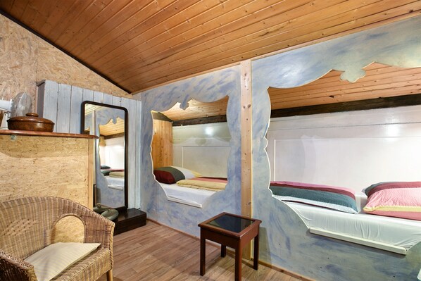 A room with wooden ceiling, bunk beds, a wicker chair, a small table, and a mirror.