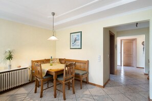 A dining area with a wooden table and chairs, a radiator, a framed picture on the wall, and a hallway leading to another room.