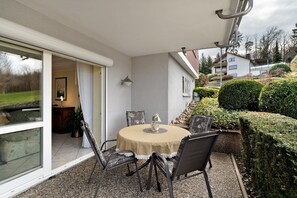 Eine überdachte Terrasse mit rundem Tisch und Stühlen, eine Glastür zu einem Innenraum und Blick auf ein Haus und einen Garten.