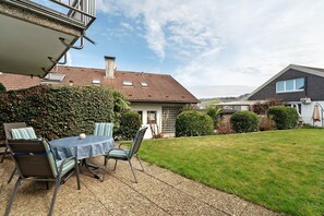 A patio with a table and chairs, a hedge, and a house with a tiled roof.