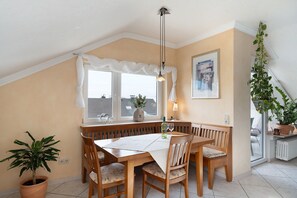 A dining area with a table, chairs, a bench, a window with curtains, a hanging plant, and a radiator.
