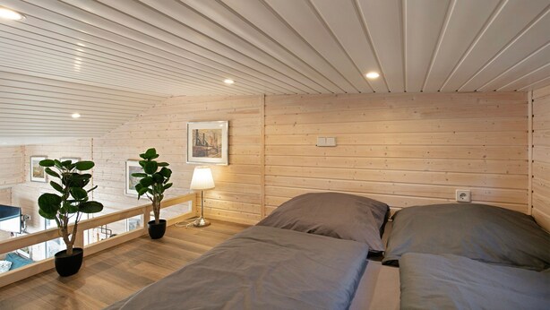 A room with a sloped ceiling, wooden walls, a bed with grey bedding, a bedside table with a lamp, and two potted plants. - Tiny Home 'Tiny House Nr 3' with private terrace, Wi-Fi and air conditioning (Schwielowsee)