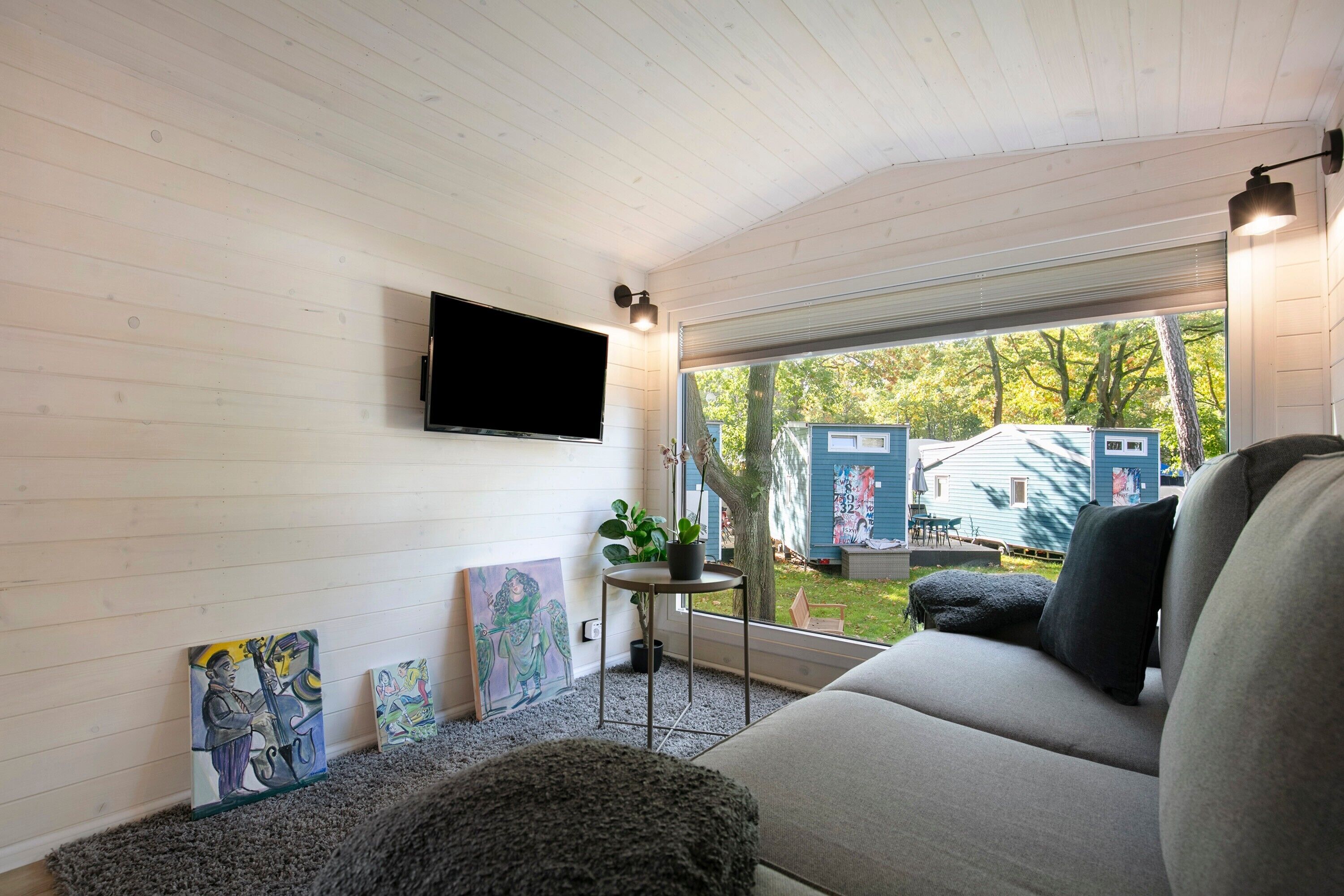 A room with a flat-screen TV mounted on the wall, a grey sofa, a small round table with a plant, and artwork on the walls.