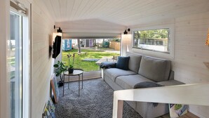 A cozy room with a sofa, a small table, a potted plant, and a window overlooking a yard. - Tiny Home 'Tiny House Nr 4' with private terrace, Wi-Fi and air conditioning (Schwielowsee)