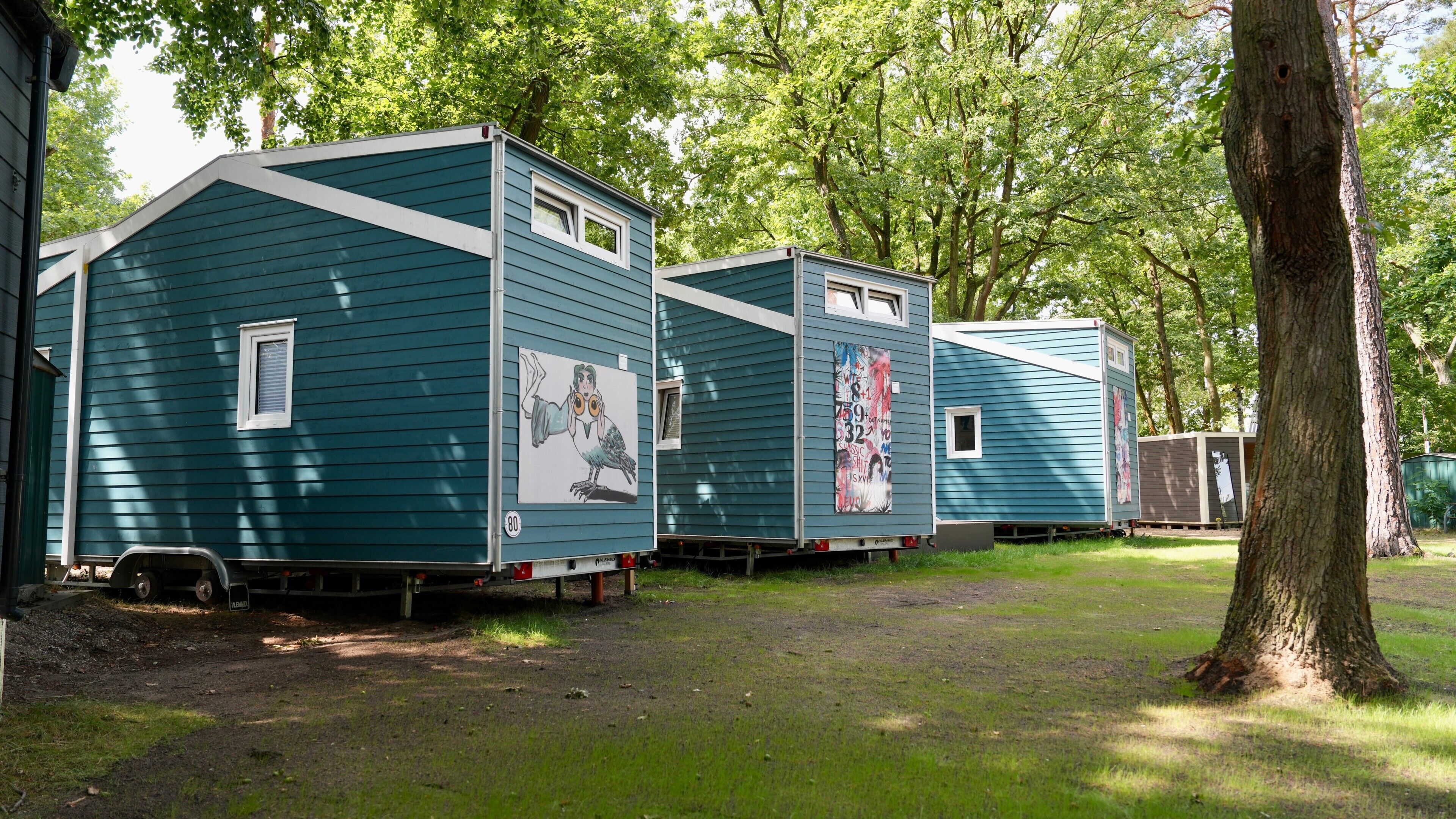 A row of blue mobile homes with artwork on the sides, parked in a grassy area with trees in the background.