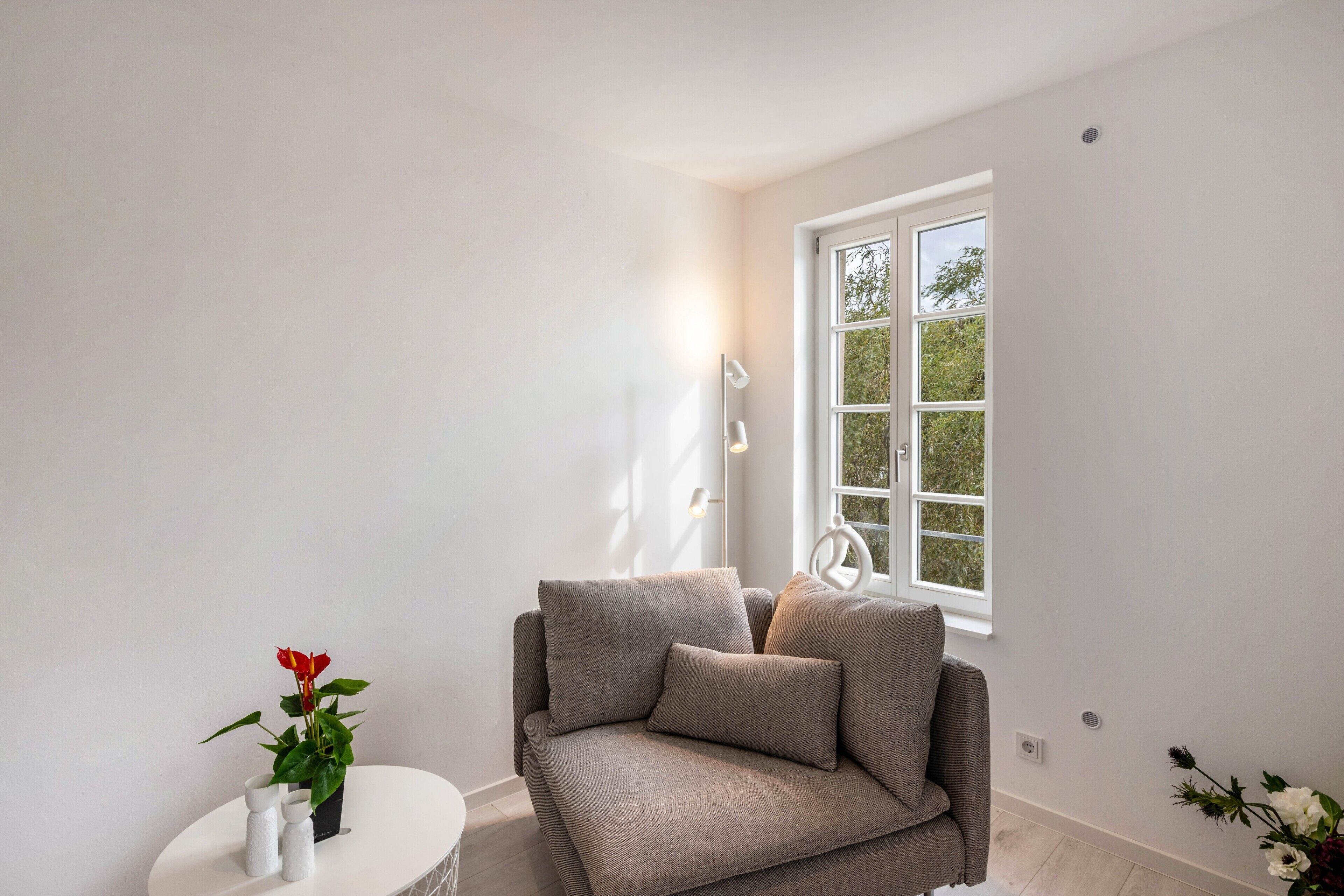 Un salon minimaliste avec un canapé gris, une petite table d’appoint blanche et une fenêtre donnant sur un paysage verdoyant.