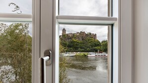 Vista desde una ventana de un castillo junto a un río, con barcos y vegetación.