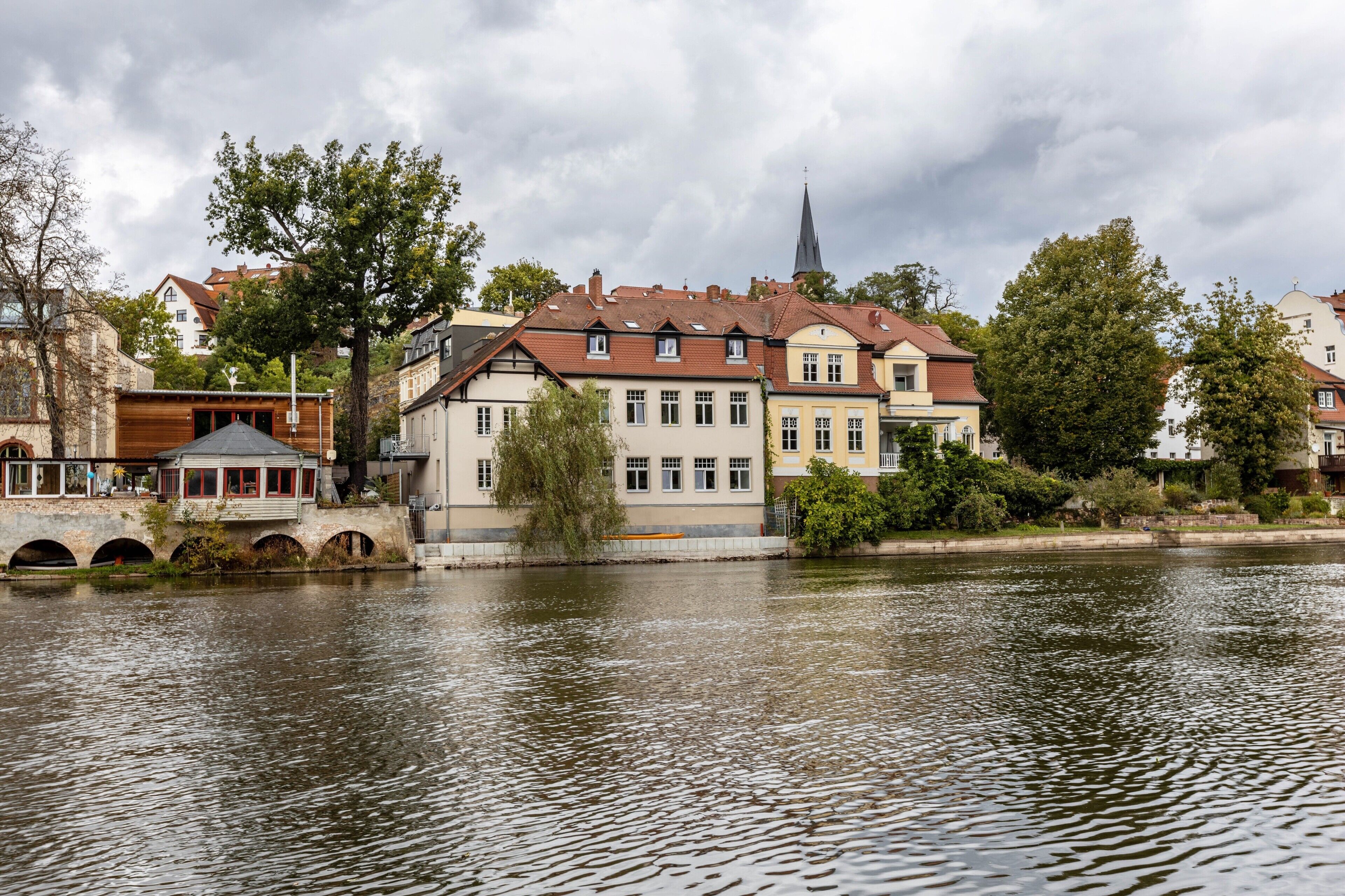 Un edificio lungo il fiume con uno stile architettonico tradizionale, una guglia di chiesa sullo sfondo e un ponte ad archi.