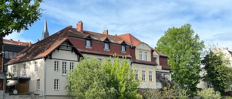A historic building with a river in front, a church spire in the background, and lush greenery.