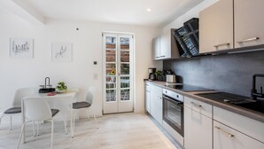 Cocina moderna con armarios blancos, encimera negra y horno empotrado. Cuenta con una zona de comedor con mesa y sillas blancas, y una puerta que da a un espacio exterior.