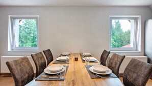 Een eetkamer met een houten tafel voor zes personen, aan weerszijden van twee ramen met uitzicht op bomen.